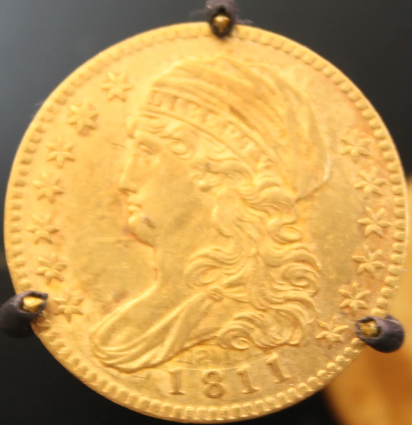 Obverse of an 1811 half eagle held in display clips, Liberty facing left in a soft cloth cap with LIBERTY lettered on the headband