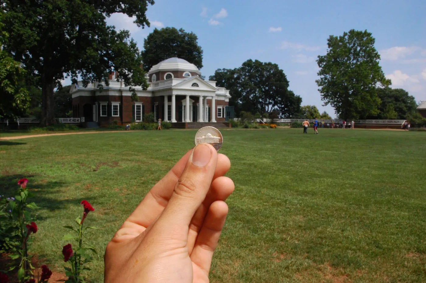 A Jefferson nickel held reverse-outward in front of the east front of Monticello, the coin's building matching the portico and dome behind it