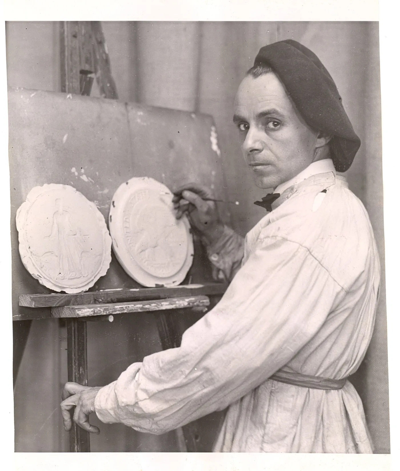 Anthony de Francisci in a studio smock working on two large circular plaster models, the left showing a standing figure and the right lettered UNITED STATES