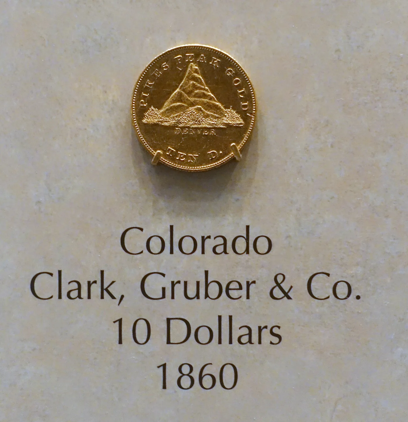 An 1860 Clark, Gruber and Company ten dollar gold coin on museum display, its obverse showing a stylised Pikes Peak with the legend PIKES PEAK GOLD and TEN D., above a label reading Colorado, Clark, Gruber and Co., 10 Dollars, 1860