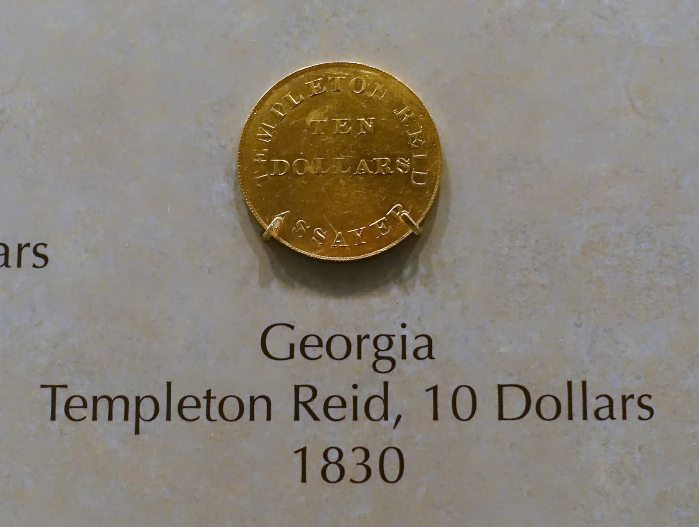 An 1830 Templeton Reid ten dollar gold coin on museum display, lettered TEMPLETON REID ASSAYER around TEN DOLLARS, above a label reading Georgia, Templeton Reid, 10 Dollars, 1830